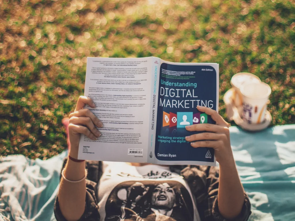 Man lying on the ground in the park with a book about digital marketing in his hands, enjoying a sunny day with trees and greenery in the background.