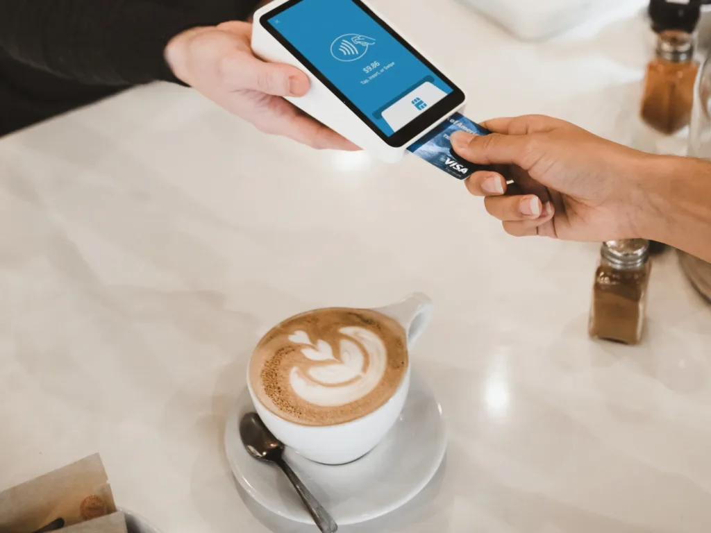 A person at a coffee shop checkout counter, holding a credit card and looking at a screen while a barista hands over a freshly brewed cappuccino. The coffee shop environment is visible in the background with other patrons and decor elements. The transaction showcases a cashless payment method in a modern setting.