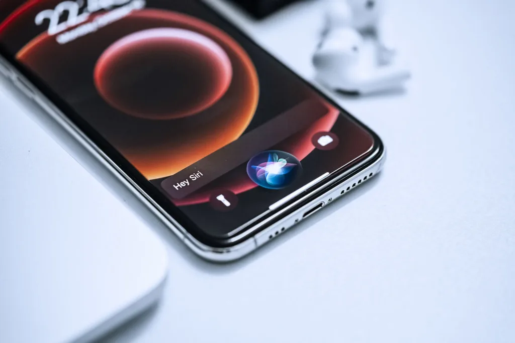 An iPhone sits on a desk with Siri, the virtual assistant, displayed on the screen. Siri responds to a user's question, displayed in a chat bubble on the phone's screen. The background features various office supplies and a computer monitor in the distance.