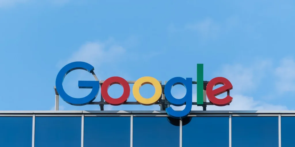 The iconic Google logo sign in bright, primary colors against a clear blue sky background, represents the well-known search engine and technology company.