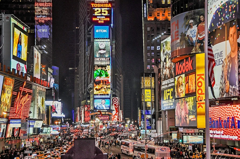 This image shows the bustling Times Square at night, brightly lit by numerous colorful billboards and neon signs advertising various brands and shows. The crowded streets are filled with a plethora of yellow taxis and pedestrians, showcasing the vibrant energy of New York City. The scene captures the essence of urban life, commercialism, and the iconic cultural landmark that is known worldwide.